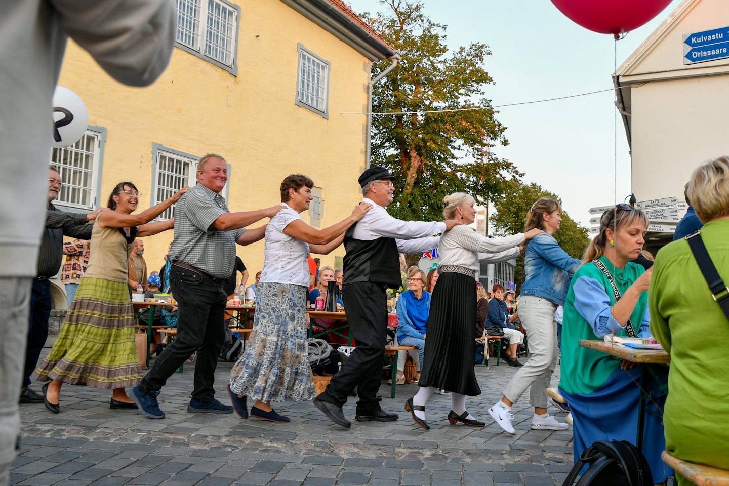 Menschen halten sich an den Schultern, während sie auf einer Kopfsteinpflasterstraße Square Dance tanzen.